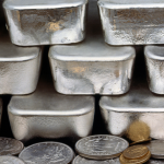 A stack of silver bullion bars atop a pile of gold and silver coins.