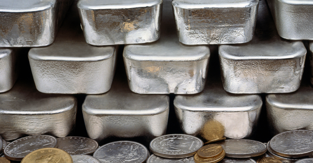 A stack of silver bullion bars atop a pile of gold and silver coins.